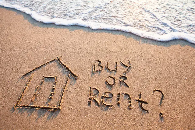 House drawn in the sand on the beach and the words "buy or rent?"